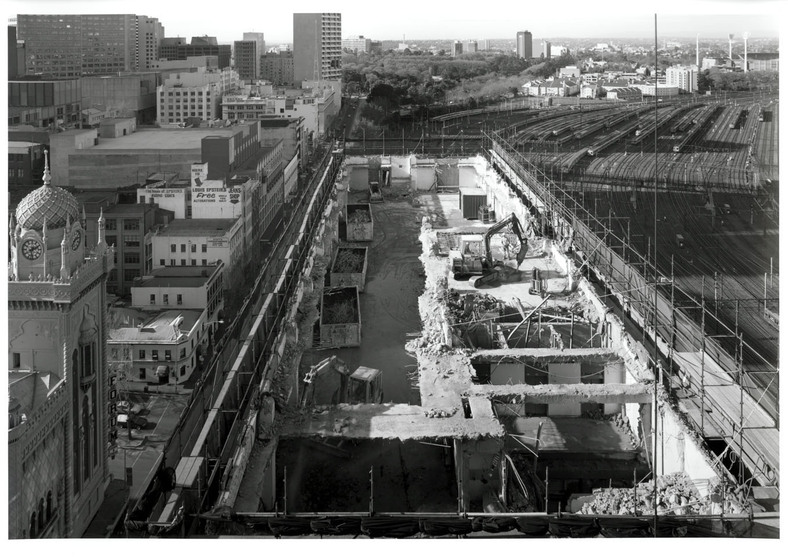 Black and white photograph of a partially demolished building from a high vantage point. Railyards beyond.