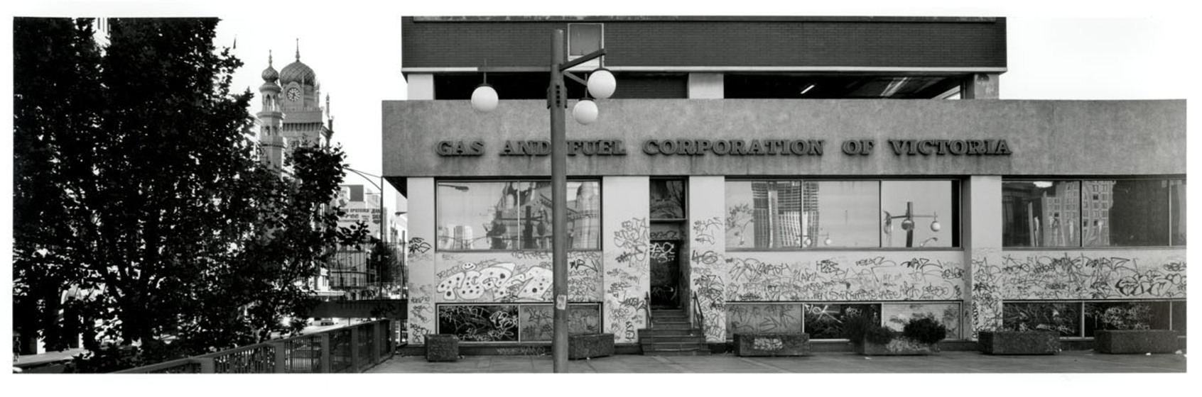 Black and white photograph of the front of a modernist building covered in graffiti with a Moorish-style clock tower to the left in the distance. Sign on the building says, 'Gas and Fuel Corporation of Victoria'.