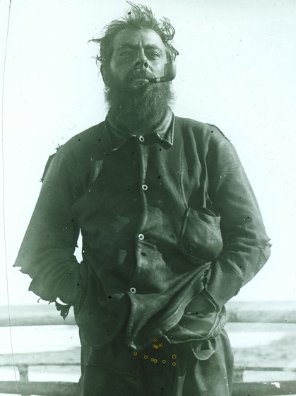 Black and white three quarter photo portrait of a man with wind blown hair, a beard and his hands in the pockets of his leather jacket smoking a pipe.