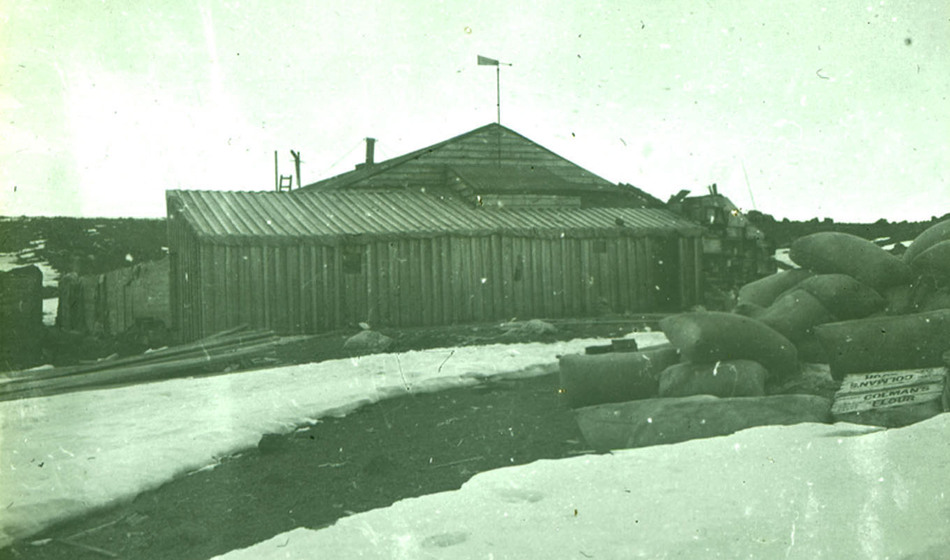 Green tinted black and white photograph of wooden hut amongst ice. Pile of full sacks in foreground.