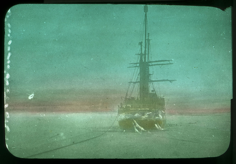 Black and white hand-coloured photograph of a square-rigged sailing ship surrounded by ice.