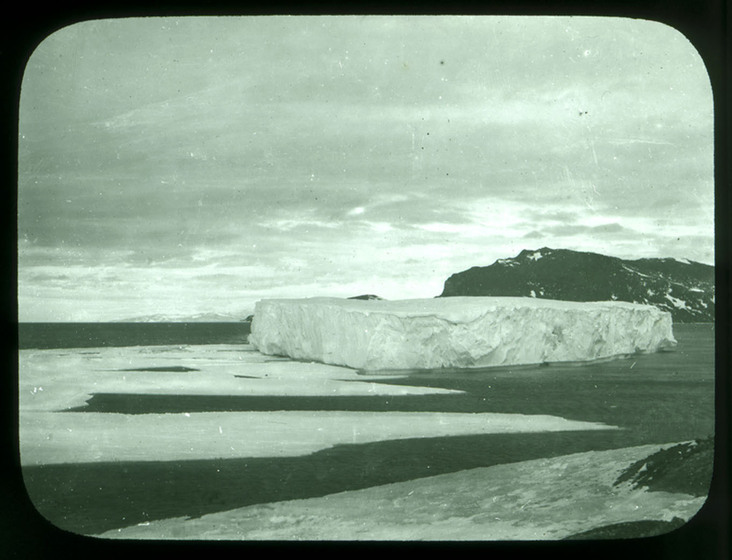 Black and white photograph of a grounded iceberg, mountain behind.