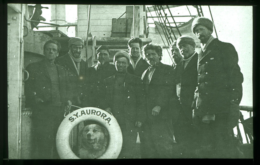 Black and white photograph of nine men standing on the deck of a sailing ship with a life preserver with 'S.Y. Aurora' on it. A dog looks through the life preserver.