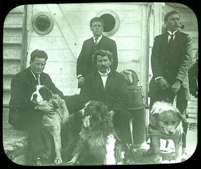 Black and white photograph of four men in Edwardian suits holding three large dogs outside a wooden structure.