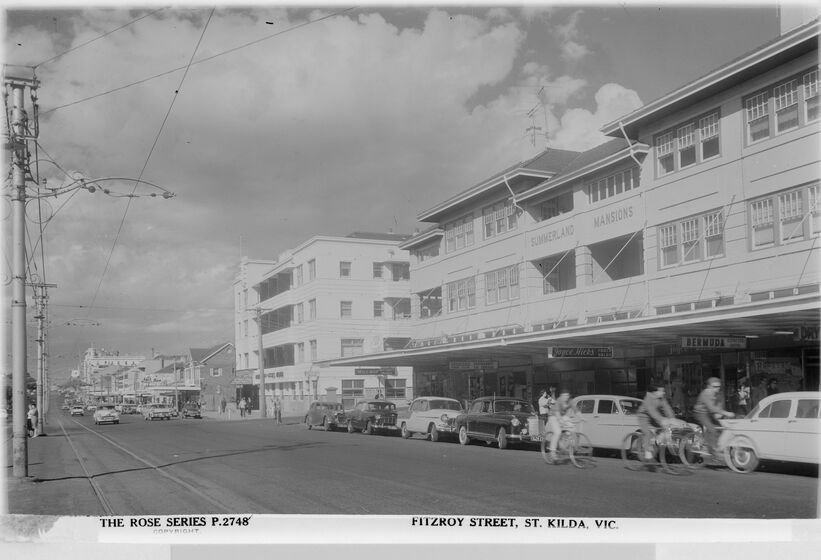 Black and white photo postcard looking along a suburban shopping strip in the 1950s with a 1920s hotel and apartment block, cars parked along the street and three people riding bicycles.