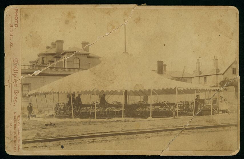 Black and white photograph of a three men in Victorian clothes riding bicycles on a bicycle carousel under a white tent.