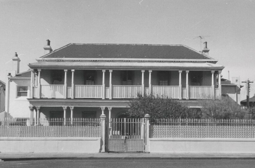 Black and white photograph of a two-storey Victorian mansion behind an iron fence with verandahs on both levels.