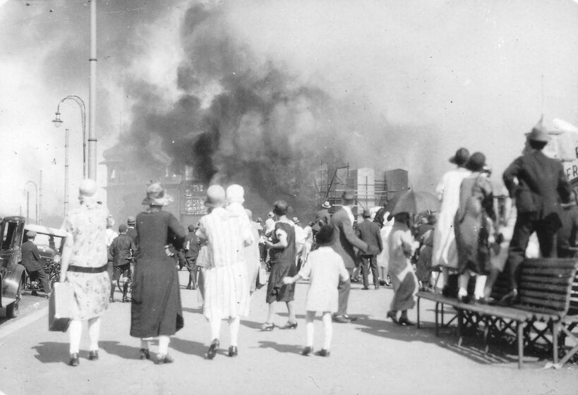 Black and white photograph of a crowd in 1920s attire watching a building engulfed in fire.