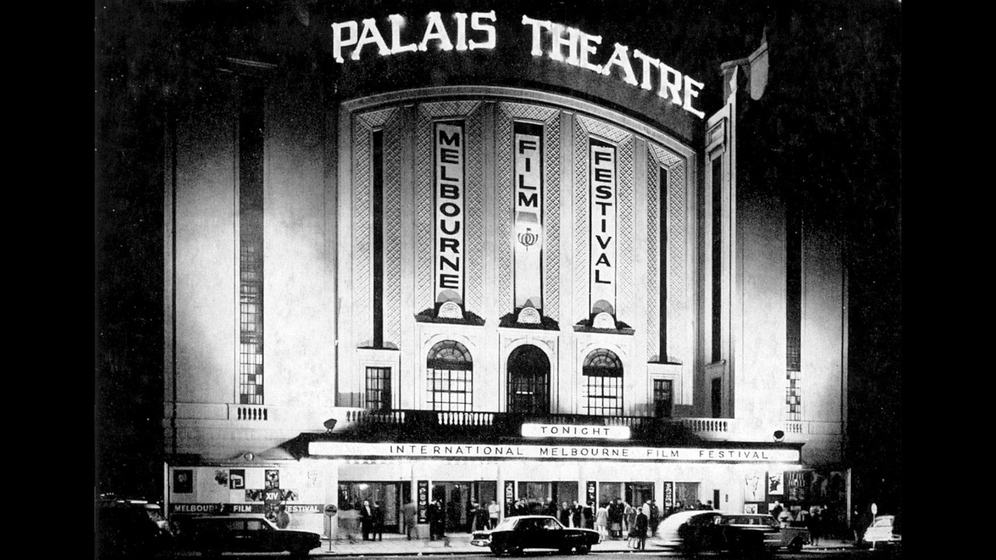 Black and white photograph of the flood lit façade of an Art Deco cinema at night with the sign, 'Palais Theatre'.