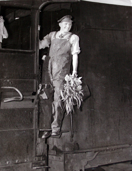 Black and white photograph of a man standing on the ledge outside of a steam train cabin. He is wearing overalls, a cap and holding onto a bunch of eucalyptus leaves and flowering buds.