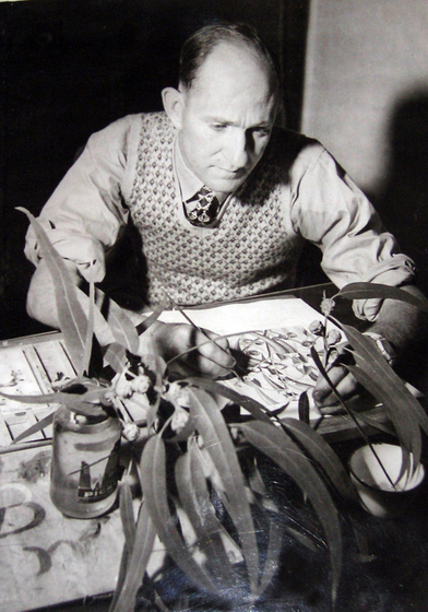 An older man wearing a patterned vest is poised over a table. He has a paint brush in one hand and is looking at a series of gum leaves resting in a jar of water. A half finished painting is on the table in front of him.