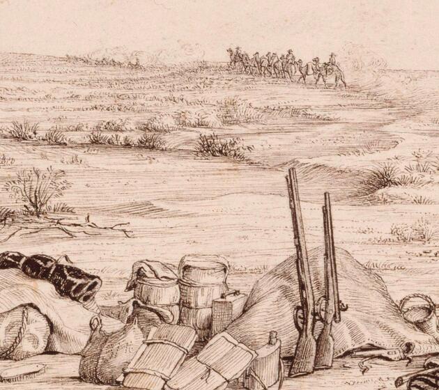 A large pile of equipment, including boxes, water carriers, bags and rifles lie on the ground. There is minimal landscape, indicating the scene is in the desert.
