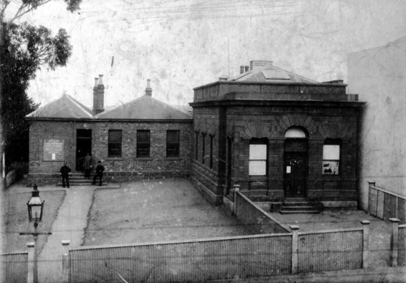 Black and white image of two stone buildings, side by side. They are set back from the road, with a short tin fence at the front of the yard. Two men stand in front of one of the stone buildings, they look to be in police uniform.
