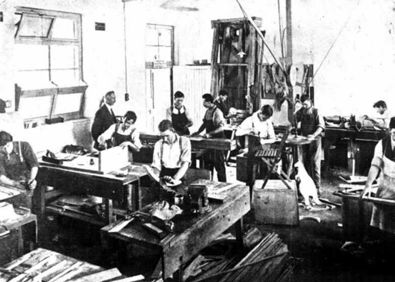 Group of men in a workshop, all at individual benches working on various projects such as sanding, plaining or hammering.