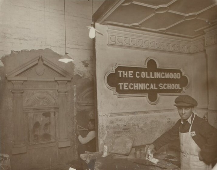 Man standing with an apron in front of a sign that reads 'The Collingwood Technical School'. He is holding a tool in one hand and staring at the camera. In the background an man is working on plastering a decorative feature onto a wall.