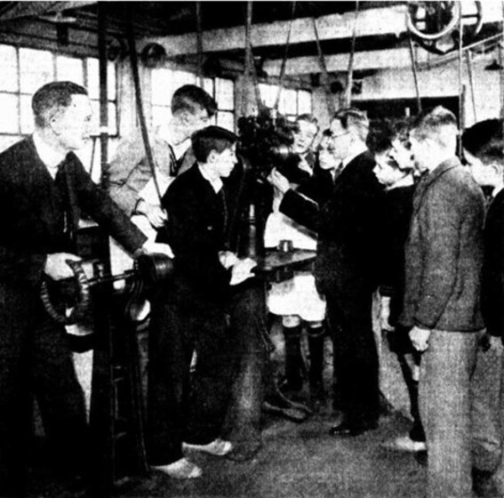 A group of young men stand in a workshop, all staring at a man in the centre of the group who is holding a new piece of machinery.