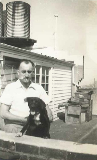 In the foreground a man stands behind a waist high brick fence, he is helping a dog stand up against the fence. He has a cigarette in his mouth. He is standing in front of the side of a weather board house with a cylinder iron tank on the roof.