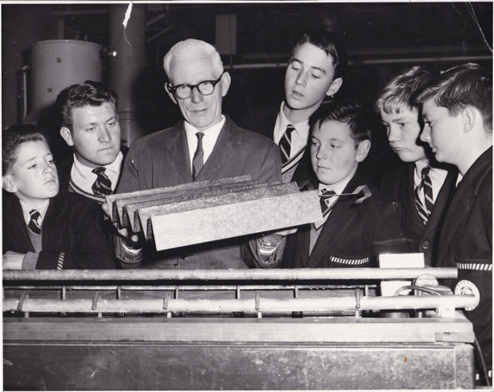 An older man stands in the centre of a group of boys in school uniform. He is holding a piece of equipment and the entire group is staring directly at it.