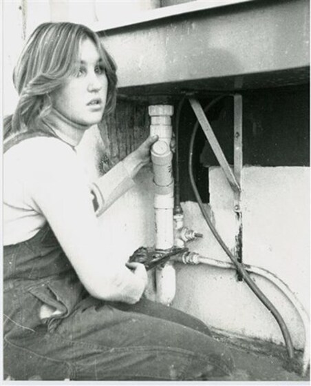 Black and white image of a woman kneeling on the ground in front of a sink pipe. She is maneuvering a wrench on one of the pipes and looking off into the distance.
