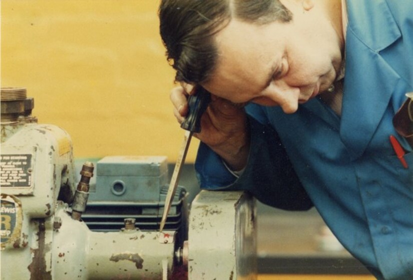 Man in a boiler suits leans over a piece of mechanical equipment, listening via a screwdriver to hear the inner workings of the machine.