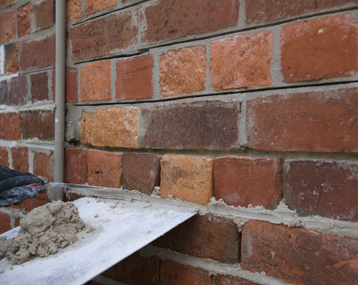 Colour photograph of an old brick wall with grey mortar on a trowel.