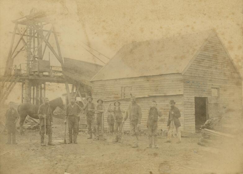 Faded and foxed black and white photograph of nine men with shovels and an unharnessed horse standing in a row in front of a colonial weatherboard building and the wooden winding gear of a mine shaft.