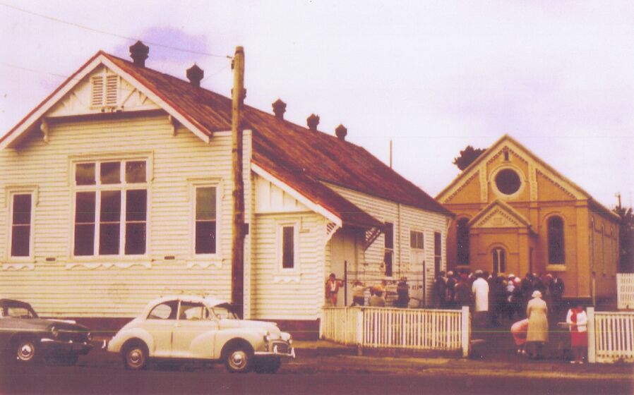 Colour photograph of a small crowd outside a Victorian brick chapel with large weatherboard hall to left.