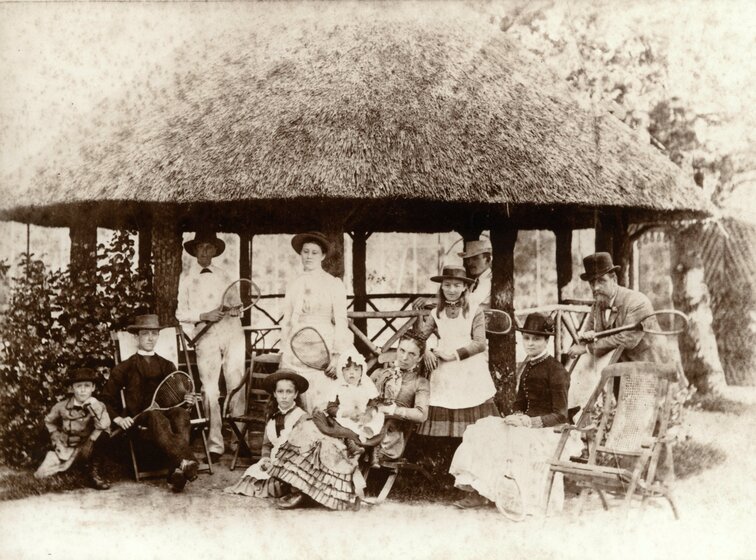 Black and white photograph of men, women and children in Victorian clothes with tennis raquets standing and sitting under a pavillion.