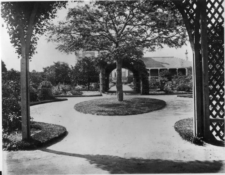 Black and white photograph of formal garden with circular white gravel path, tree in centre, house beyond, trellis to right.