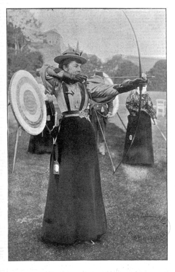 Black and white photograph of a woman in Victorian clothing standing in a garden about to shoot an arrow from an extended bow. Behind her is an archery target and other archers,