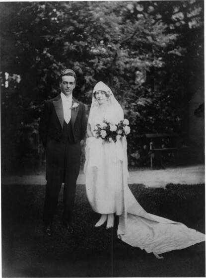 Black and white photograph of a bridal couple, groom in tails, bride in 1920s style wedding gown with long train holding large bouquet standing in a garden.