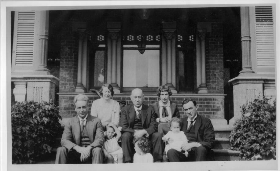 Black and white photograph of a family group of five adults and three children sitting on the entrance steps of a large house.