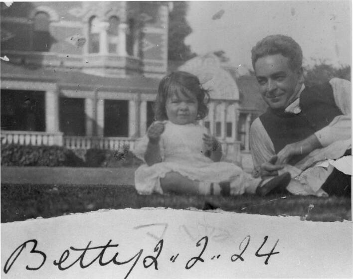 Black and white photograph of a female toddler in a white dress sitting on a lawn accompanied by a man, lying. Large house behind.