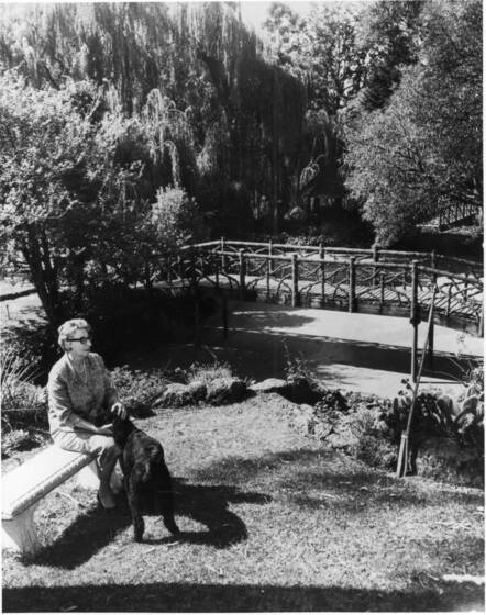 Black and white photograph of a middle-aged woman sitting on a bench in a garden with a black dog, beyond a rustic bridge over a pond and trees.