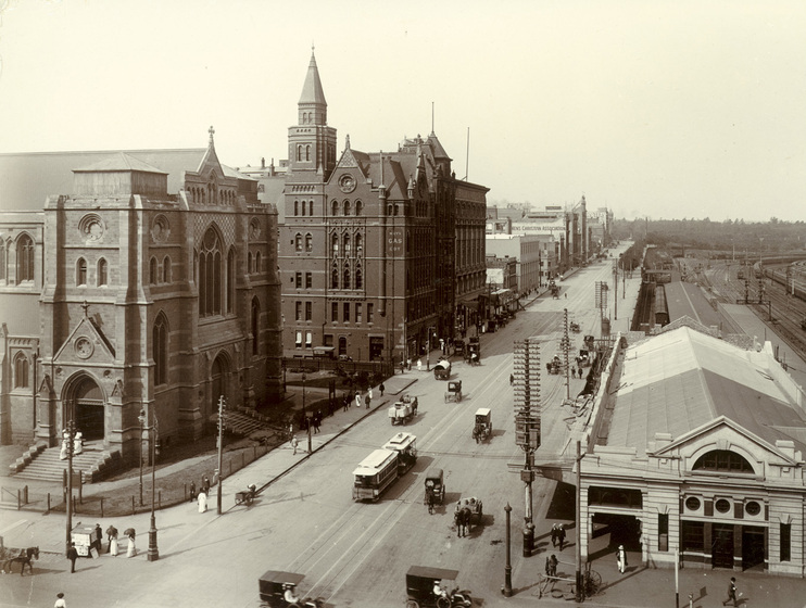 Black and white photograph looking along an Edwardian city street from a high vantage point, a Gothic Revival cathedral without towers on the left, a train station on the right, a tramcar in the street.
