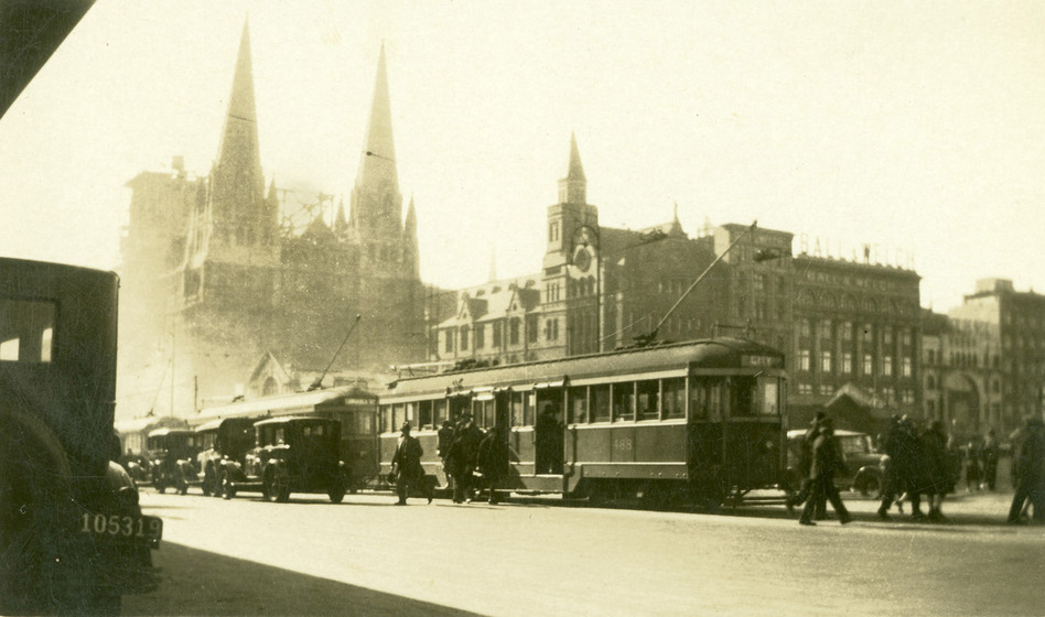 Black and white photograph of an early Twentieth Century street scene with a Gothic Revival cathedral in the background and tramcars and cars in the foreground.