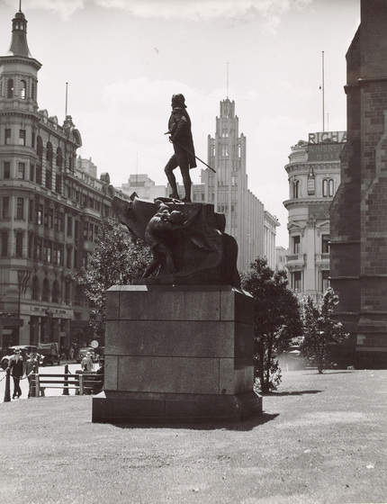 Black and white photograph of a statue of a man in Victorian dress with a naval officers hat under his arm on a plinth in a city street.