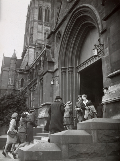Black and white photograph of people in 1940s clothing entering and leaving a Gothic Revival cathedral, including school children and a woman in military uniform.
