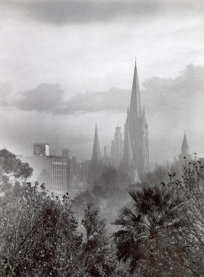 Black and white photograph of a Gothic Revival cathedral in a cityscape seen through mist. Trees in foreground.