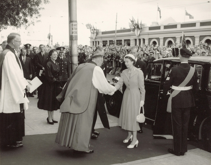 Black and white photograph of a man in archbishop's robes shaking the hand of a woman in a 1950s day dress on a pavement. Car with open door behind woman. Crowd in background.