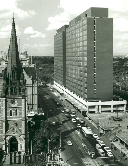 Black and white photograph of a pair of modernist multi-storey buildings from a high vantage point. Cars and a tram car in the street below. Railway lines and parkland beyond.