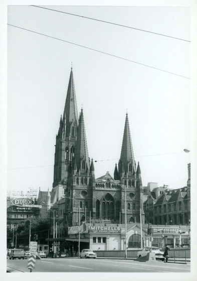 Black and white photograph of a Gothic Revival cathedral in a cityscape, partially obscured by a low building.