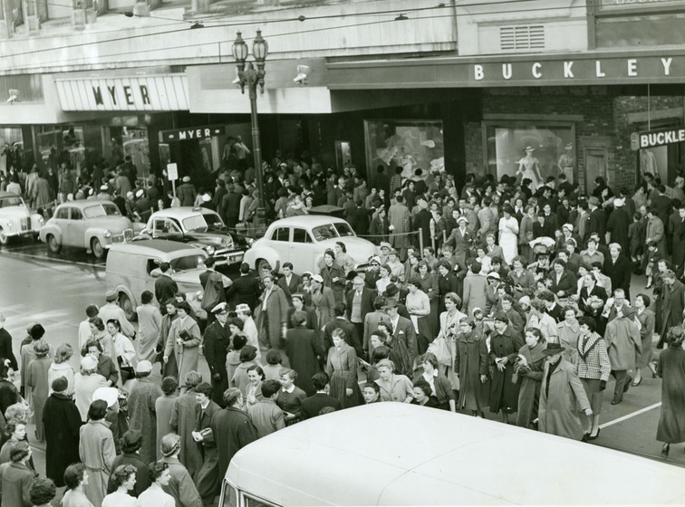 A large group of shoppers can be seen crossing a street in front of a group of shop front windows.