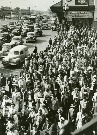 Large group of people dressed in formal attire, walk down a wide footpath. To one side of the group is an awning over the footpath, and on the other side is a busy road with lots of cars.