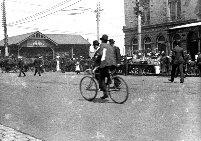 A man rides on a bicycle, dressed in a suit and hat. Behind him stands two buildings and many people walking around on the side of the street.