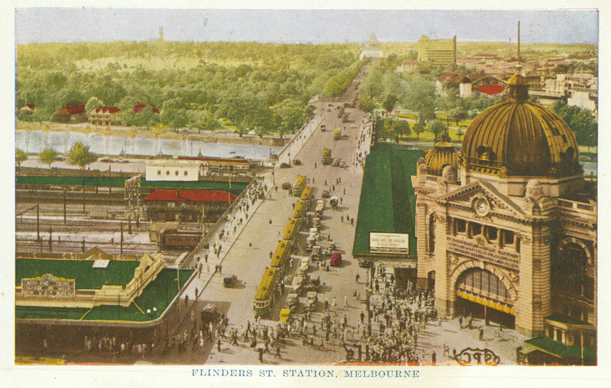 A coloured postcard of an aerial view of Flinders Street Station and the adjacent precinct. In the middle of the picture runs Swanston Street, with lots of people walking across the road and trams and cars lined up waiting to move. The river can be seen opposite the station and lots of green space where the Botanical Gardens stand.