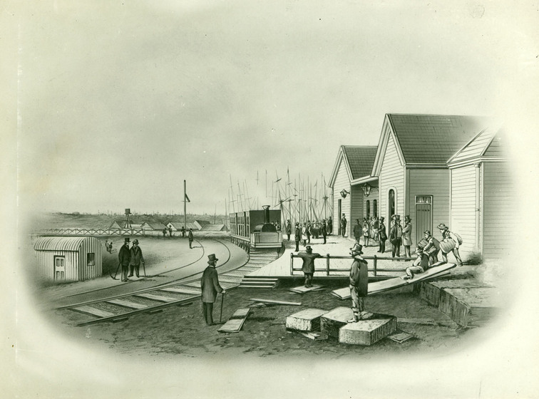 A black and white image of a railway track running past a group of wooden buildings that are most likely a train platform. Men stand around watching the train pull in, or working and lifting objects to be put on the train.