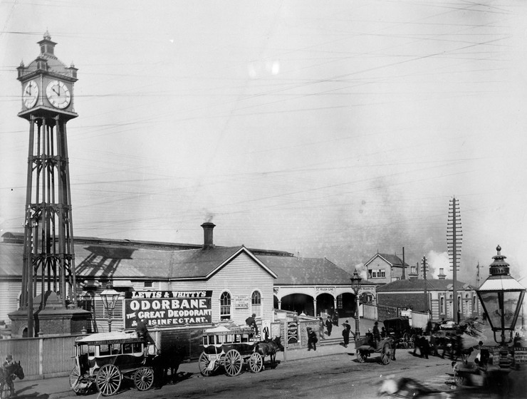 Black and white photograph of a busy streetscape. In the foreground horse carriages are lined up outside a small wooden building. To one side stands a tall metal clock, with clock faces on all four sides.
