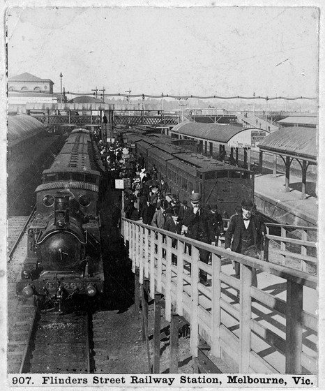 A long steam train pulls into a platform. A pedestrian ramp stands to one side of the train, and a large groups of well dressed people can be seen disembarking.