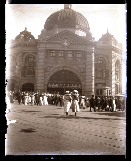 Sepia image of Flinders Street Station. The large domed roof and arched entrance ways tower over the large group of pedestrians walking past on the road at the front of the station.